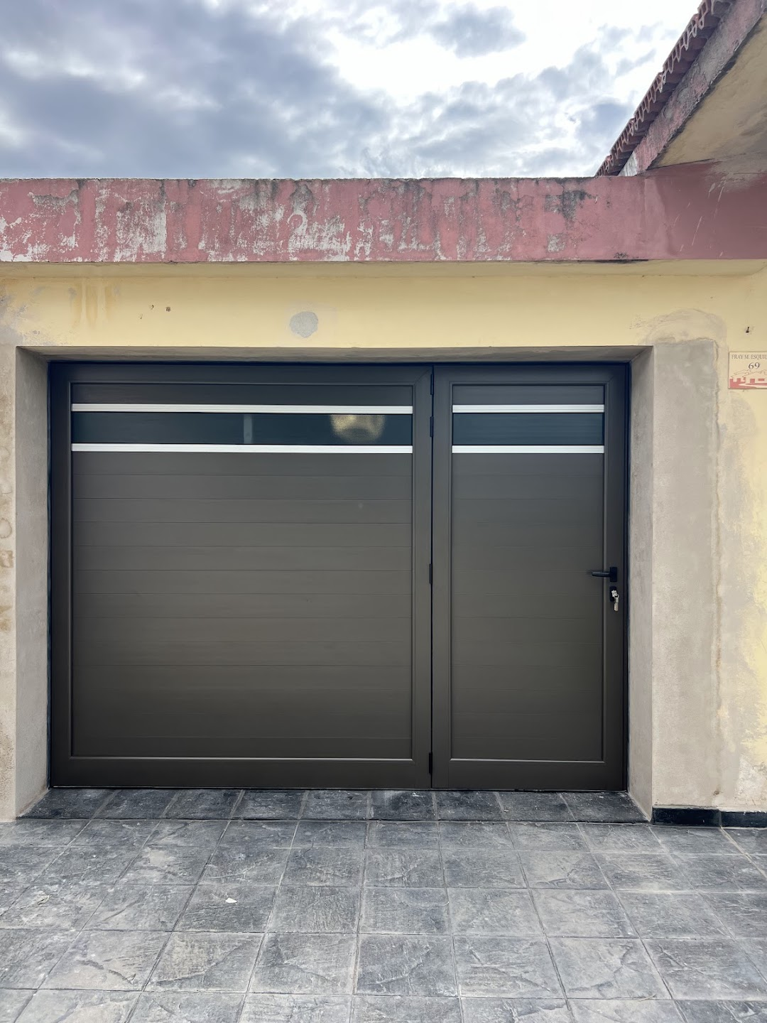 Portón oscuro con puerta lateral derecha y franja vidriada enmarcada en color anodizado natural